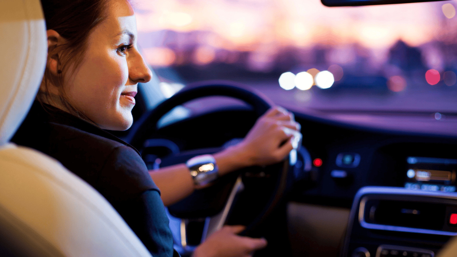 A woman is sitting in the driver's seat of a car, holding the steering wheel. The background shows blurred city lights at dusk. She is gazing forward with a slight smile, and the dashboard with various controls is visible.