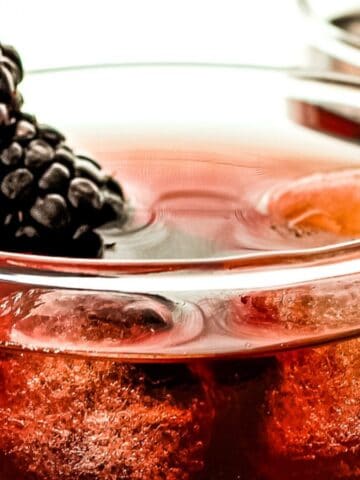 Two glasses of red berry cocktail with ice cubes, garnished with a blackberry on top, set against a white background.