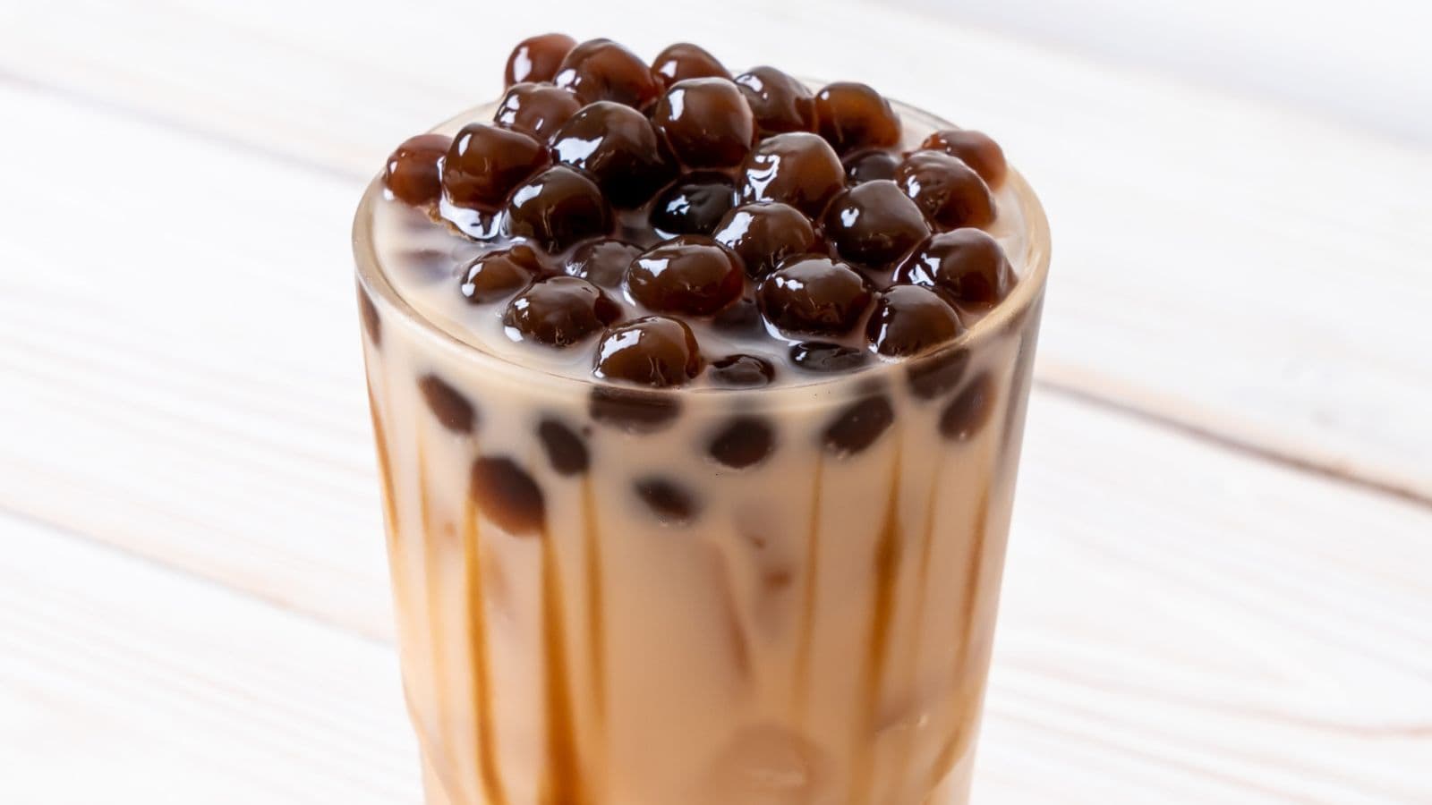 A glass of iced milk tea with a layer of chewy, black tapioca pearls at the top, sitting on a wooden table.