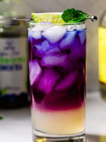 A layered cocktail with vibrant purple and white colors, served in a clear glass, garnished with a lime slice and mint leaf. a bottle and a can labeled with beverage brands are blurred in the background.