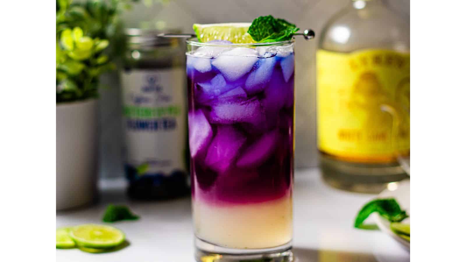 A layered cocktail with vibrant purple and white colors, served in a clear glass, garnished with a lime slice and mint leaf. a bottle and a can labeled with beverage brands are blurred in the background.