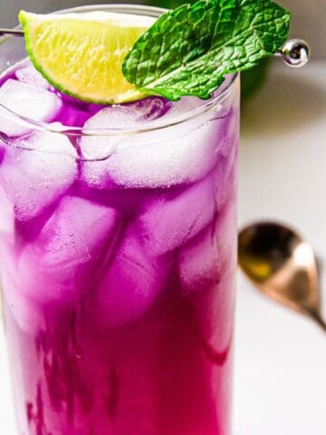 A vibrant purple cocktail in a tall glass with ice cubes, garnished with a lime wedge and a mint leaf. a white background with a cocktail shaker and lime slices scattered around.