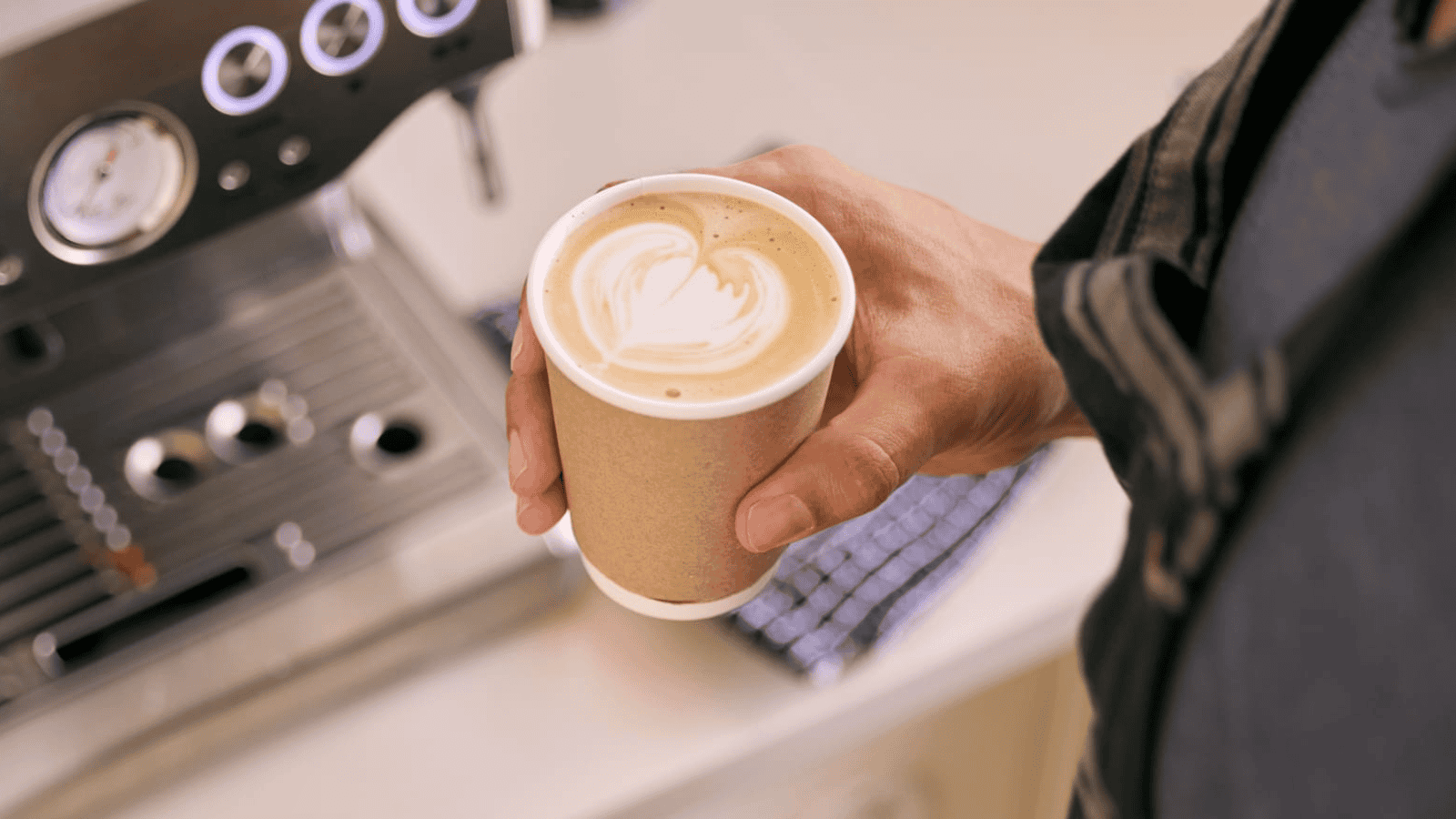 A person holds a paper cup filled with a latte, featuring a heart-shaped design in the foam. In the background, there is a coffee machine and a checkered cloth.