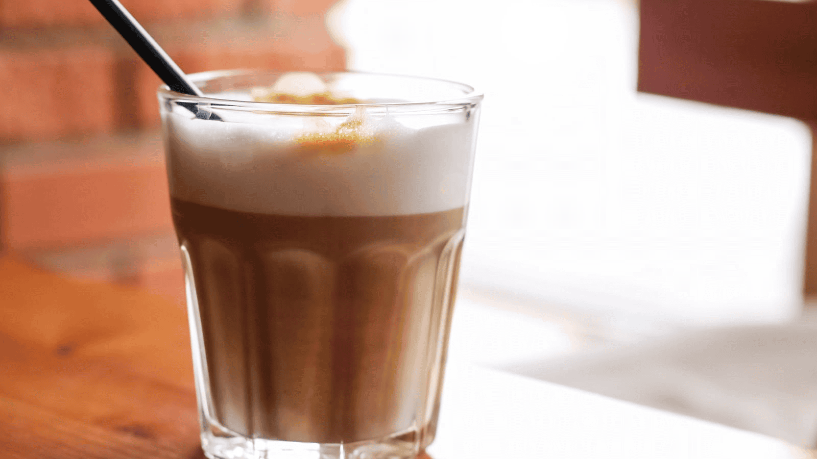 A glass of layered iced coffee with milk foam on top, placed on a wooden table. A black straw is positioned in the drink. The background is softly blurred with a hint of a brick wall and sunlight filtering through.