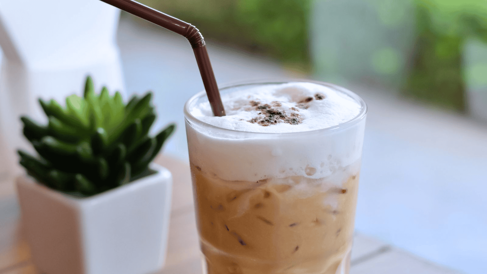 A glass of iced coffee topped with frothy milk and sprinkled with cocoa powder, featuring a brown straw. The glass is placed on a table next to a small potted succulent plant. The background is blurred, displaying greenery.