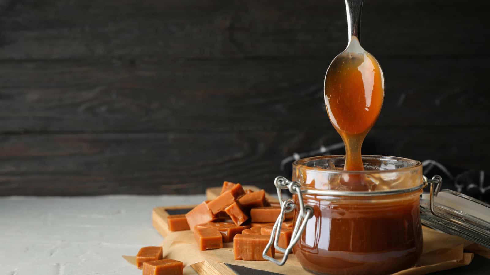 A spoon drips caramel sauce into a glass jar, surrounded by pieces of caramel candy on a wooden surface. The background consists of a dark wooden wall.