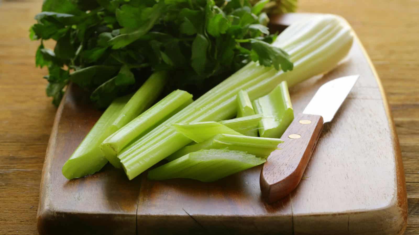 A wooden cutting board holds several stalks of celery, both whole and chopped, accompanied by a small kitchen knife with a wooden handle. A bunch of leafy green parsley is also present on the cutting board.