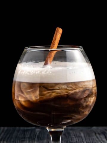 A glass of iced coffee with a frothy top and a cinnamon stick, set on a dark wooden table against a black background.