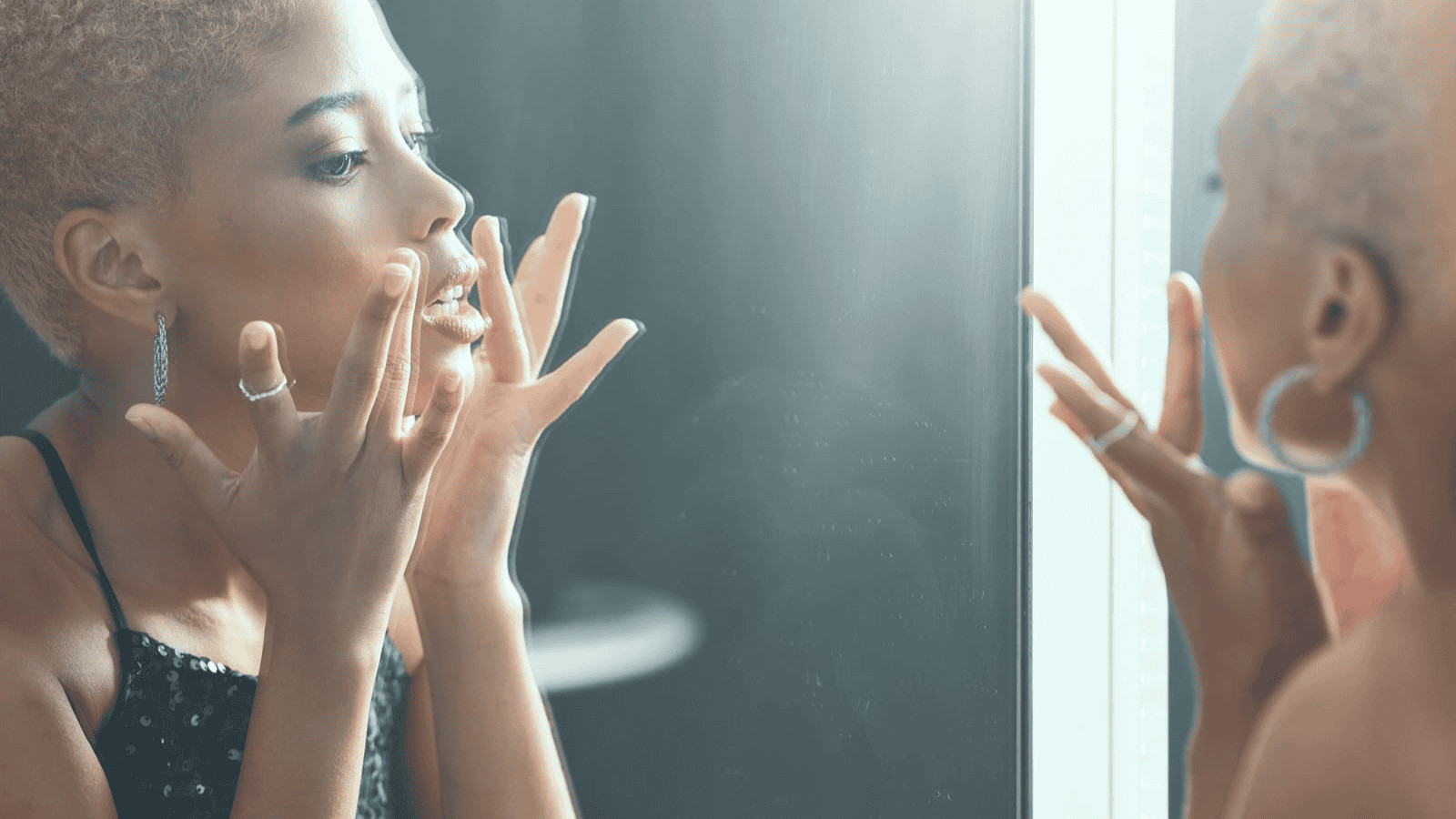 A person with short, curly hair looks intently at their reflection in a mirror, touching their face with both hands. They are wearing hoop earrings and a black, sequined top. The lighting is soft and natural.