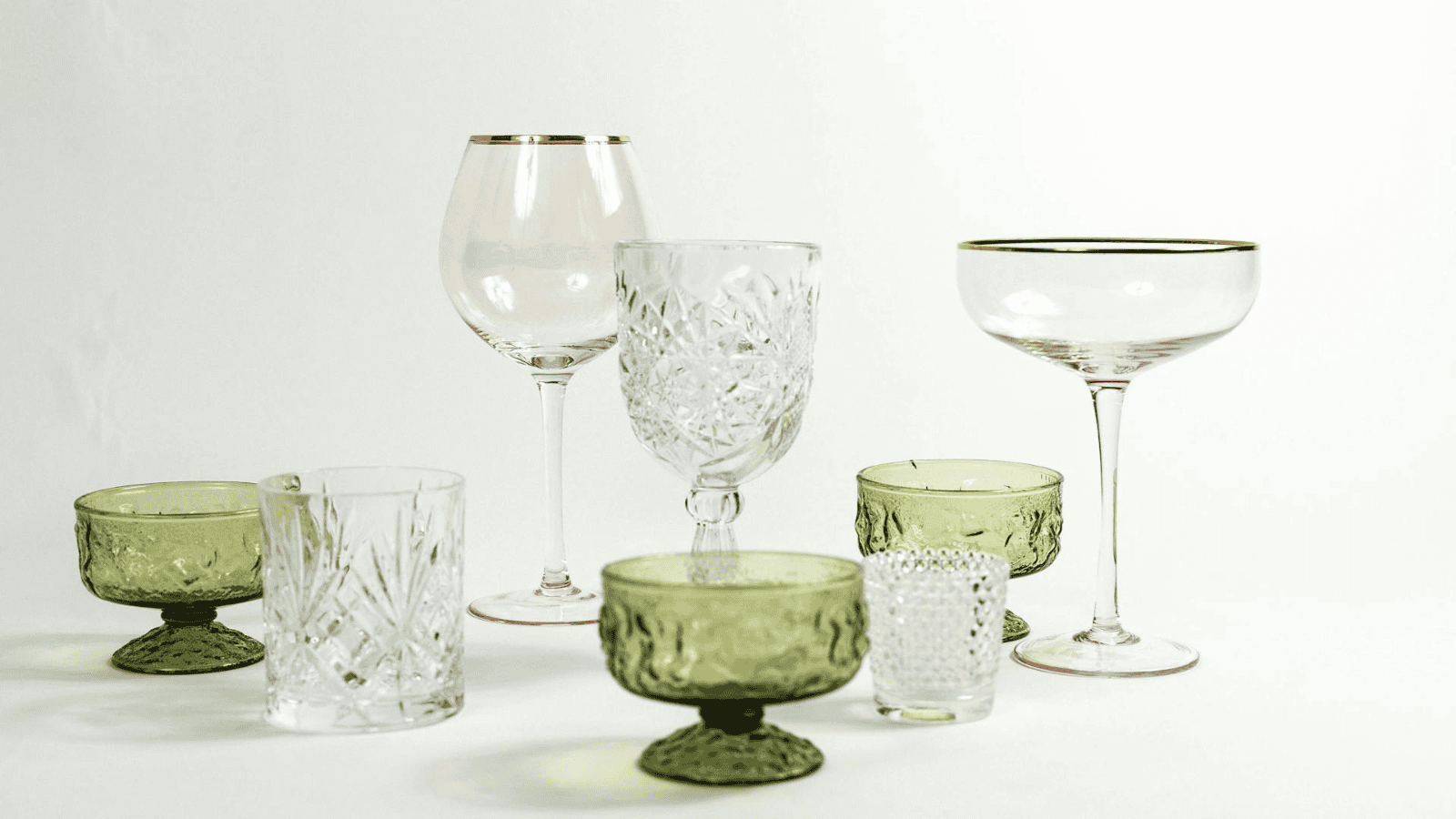 A variety of glassware, including green and clear drinking glasses and stemware, is arranged on a white background. Some glasses feature intricate patterns and textures, while others have smooth surfaces and gold rims.