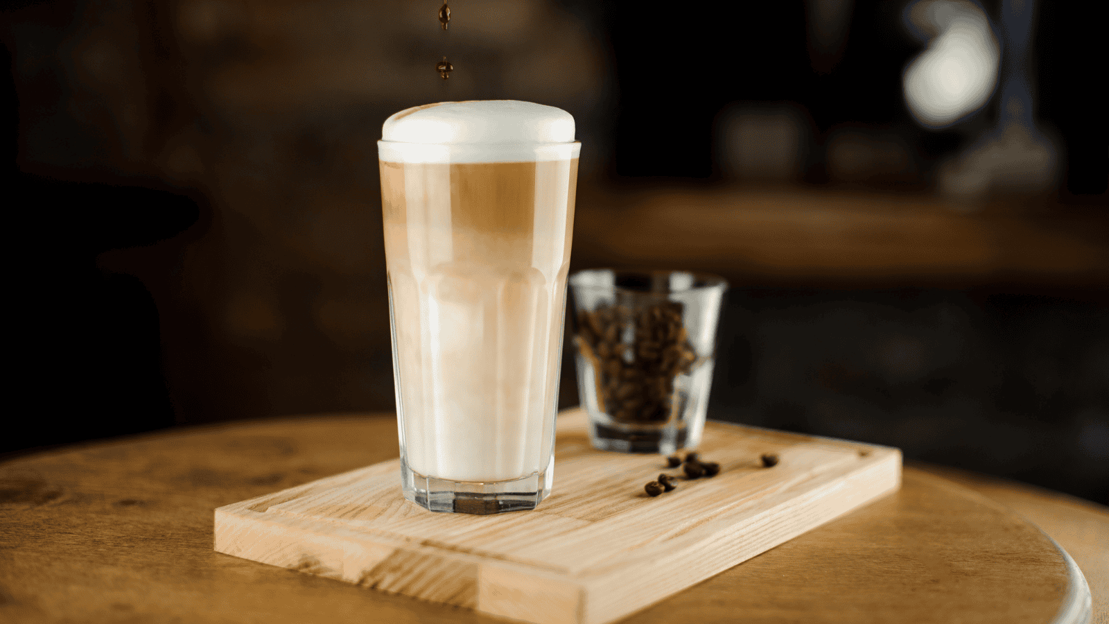 A tall glass of layered latte with foam on top sits on a wooden tray. Beside it, there is a small clear glass filled with coffee beans. The background is blurred, focusing attention on the drink and the beans.