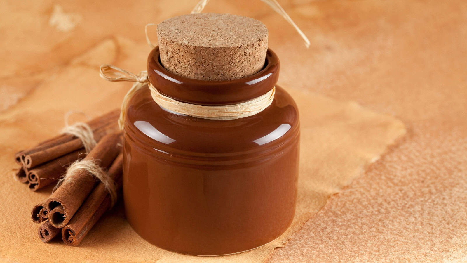 A small brown ceramic jar with a cork lid, tied with a piece of twine, is placed on a textured beige paper background. Bundles of cinnamon sticks tied with twine are situated next to the jar.