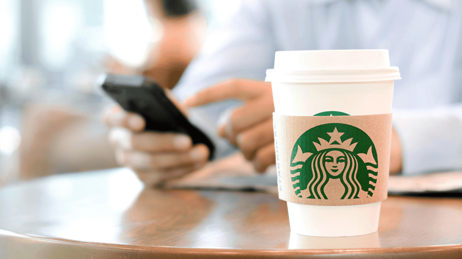 A person holding a smartphone with one hand and a newspaper in the other sits at a table. A Starbucks coffee cup with a sleeve and lid is placed on the table in the foreground.