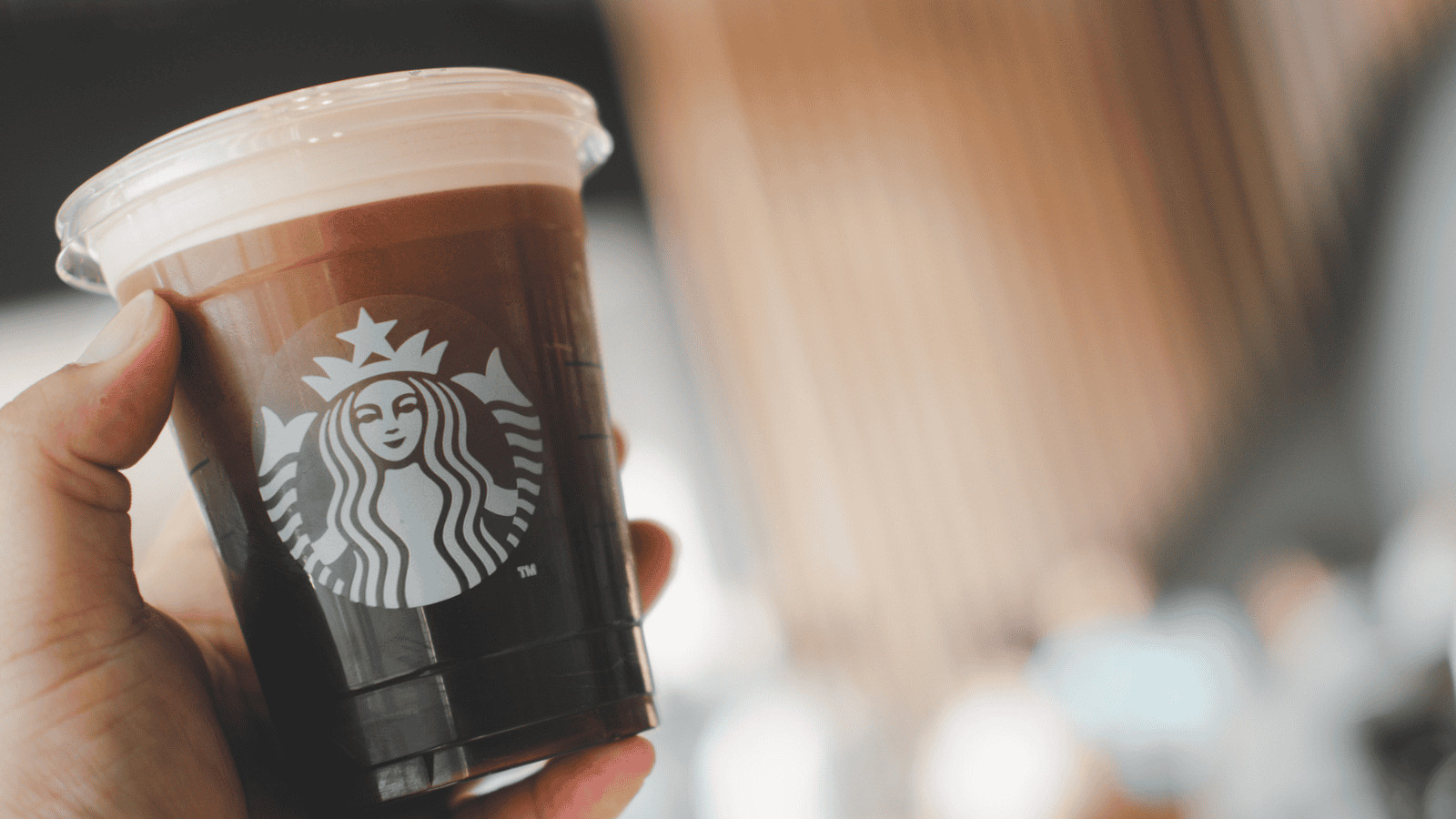 A hand holds a cold brew coffee in a clear plastic cup with the Starbucks logo. The drink has a creamy foam on top. The background is blurred, showing a lit indoor setting with indistinct figures and architectural elements.