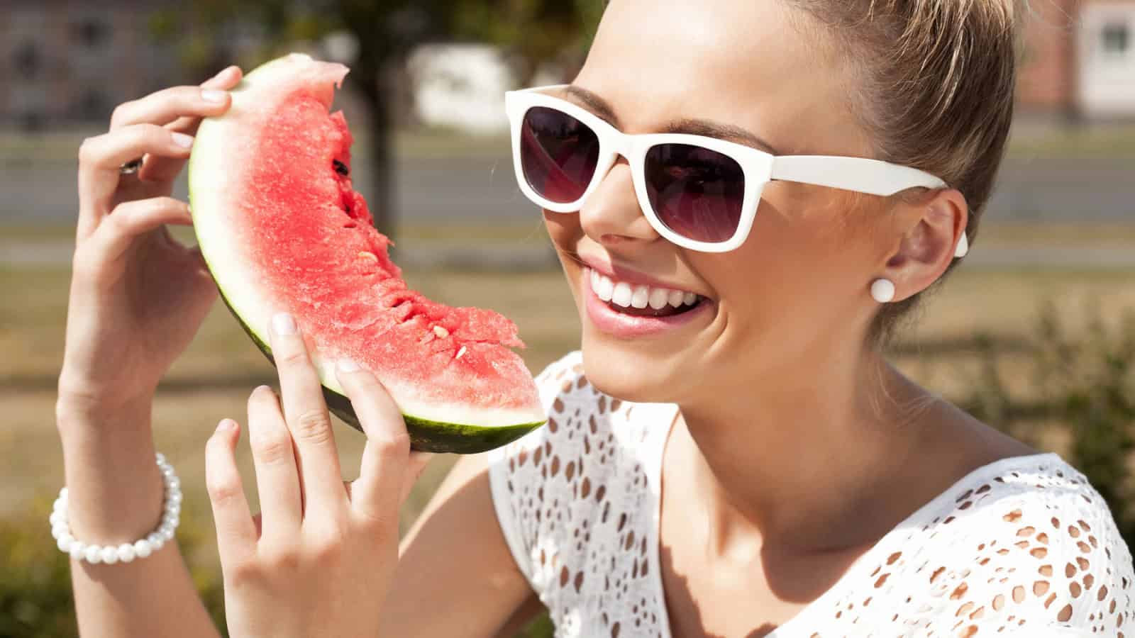 A woman wearing white sunglasses and a white top is holding a slice of watermelon close to her mouth. She is smiling and appears to be sitting outdoors on a sunny day. The background is slightly blurred with greenery and buildings visible.
