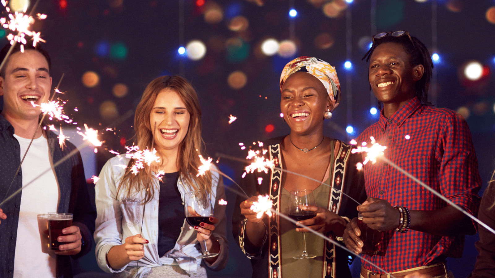 A group of four people stands together, smiling, and holding sparklers. Colorful lights in the background create a festive atmosphere. Two people are holding glasses, one with a drink in hand, and everyone appears to be enjoying the celebration.