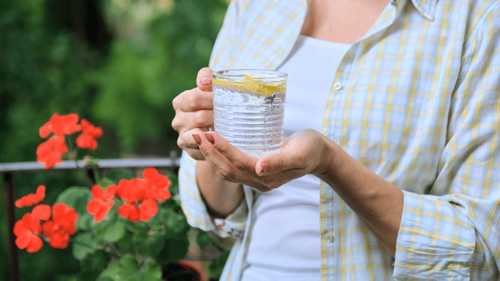 A person holds a clear glass of water with a lemon slice. The person is dressed in a white shirt and a yellow plaid button-up shirt. Red flowers are visible in the background, surrounded by green foliage.