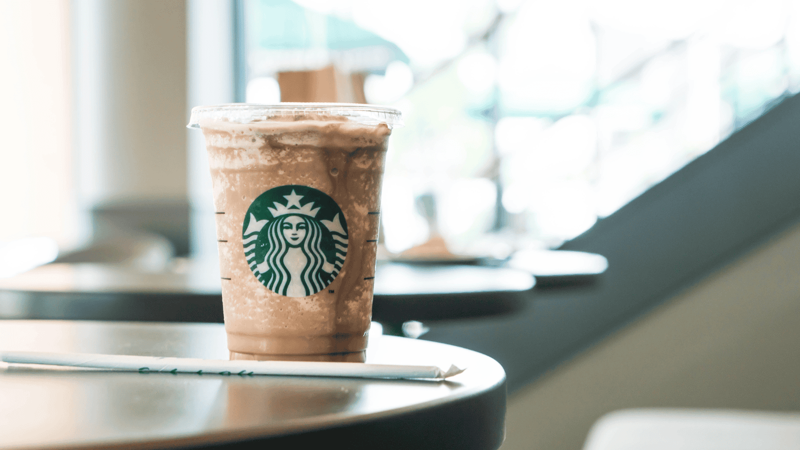 A plastic cup of a blended iced coffee drink with whipped cream topped with chocolate syrup from Starbucks is placed on a round table. A green straw wrapped in paper lies on the table in front of the cup. The background shows a blurred interior of a caf&eacute;.