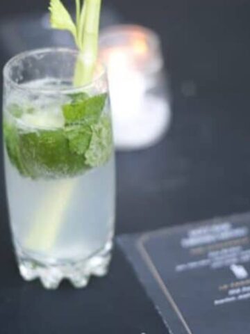 A glass of mojito with lime and mint on a dark table, next to a menu card and a lit candle, in a softly focused background.