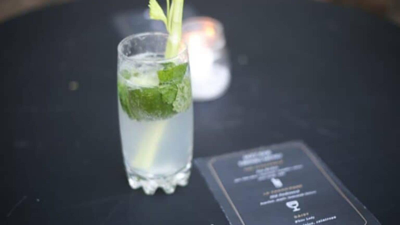 A glass of mojito with lime and mint on a dark table, next to a menu card and a lit candle, in a softly focused background.