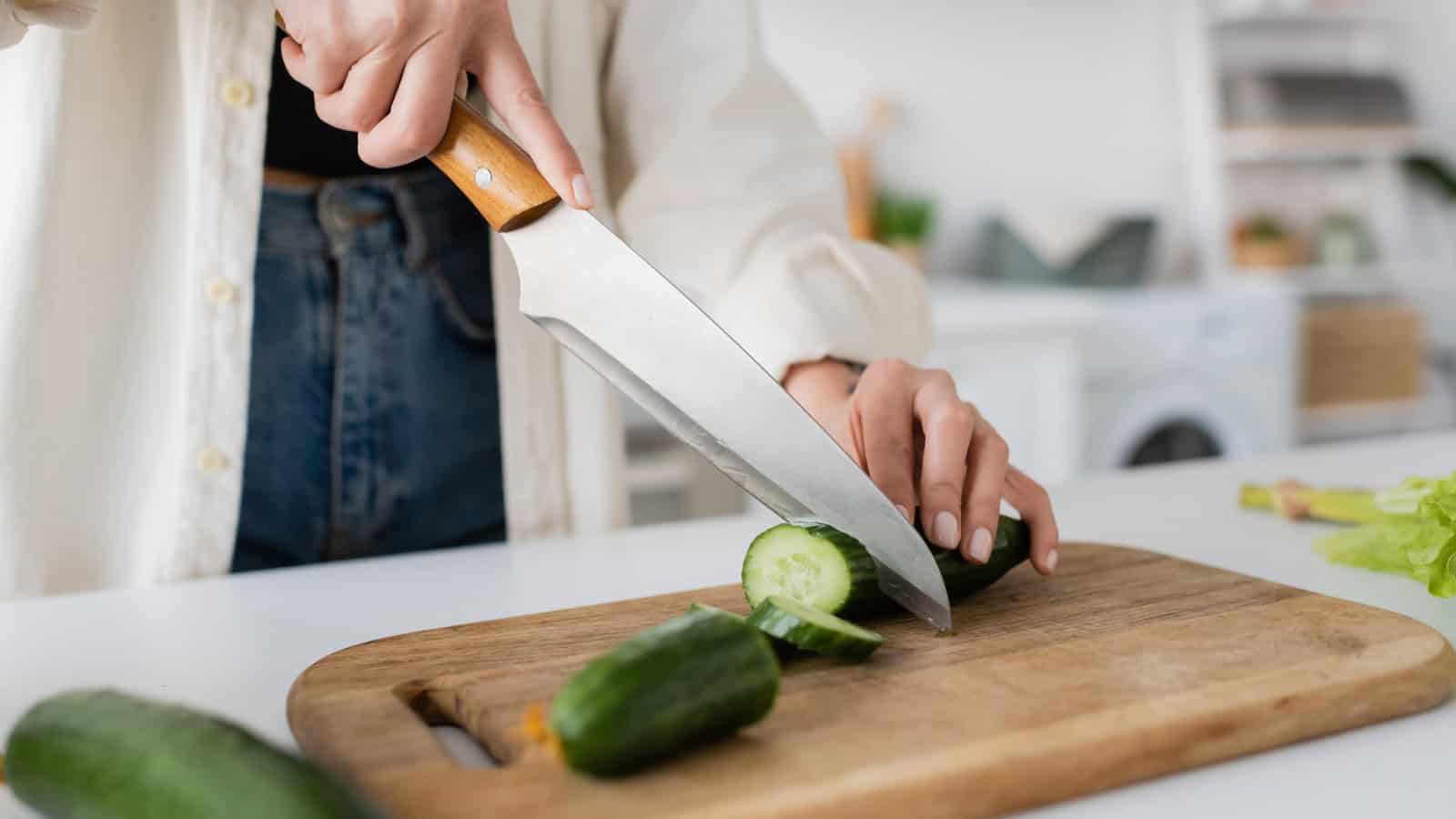 A person is slicing a cucumber on a wooden cutting board. The knife has a wooden handle, and the person is wearing jeans and a white shirt. The background shows a kitchen with various items out of focus.