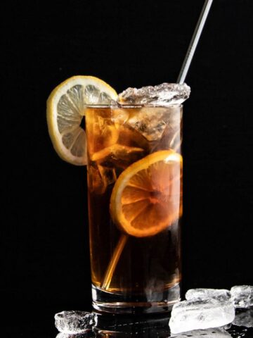 A glass of iced tea with lemon slices on a black background, accompanied by scattered ice cubes and a stirring straw.