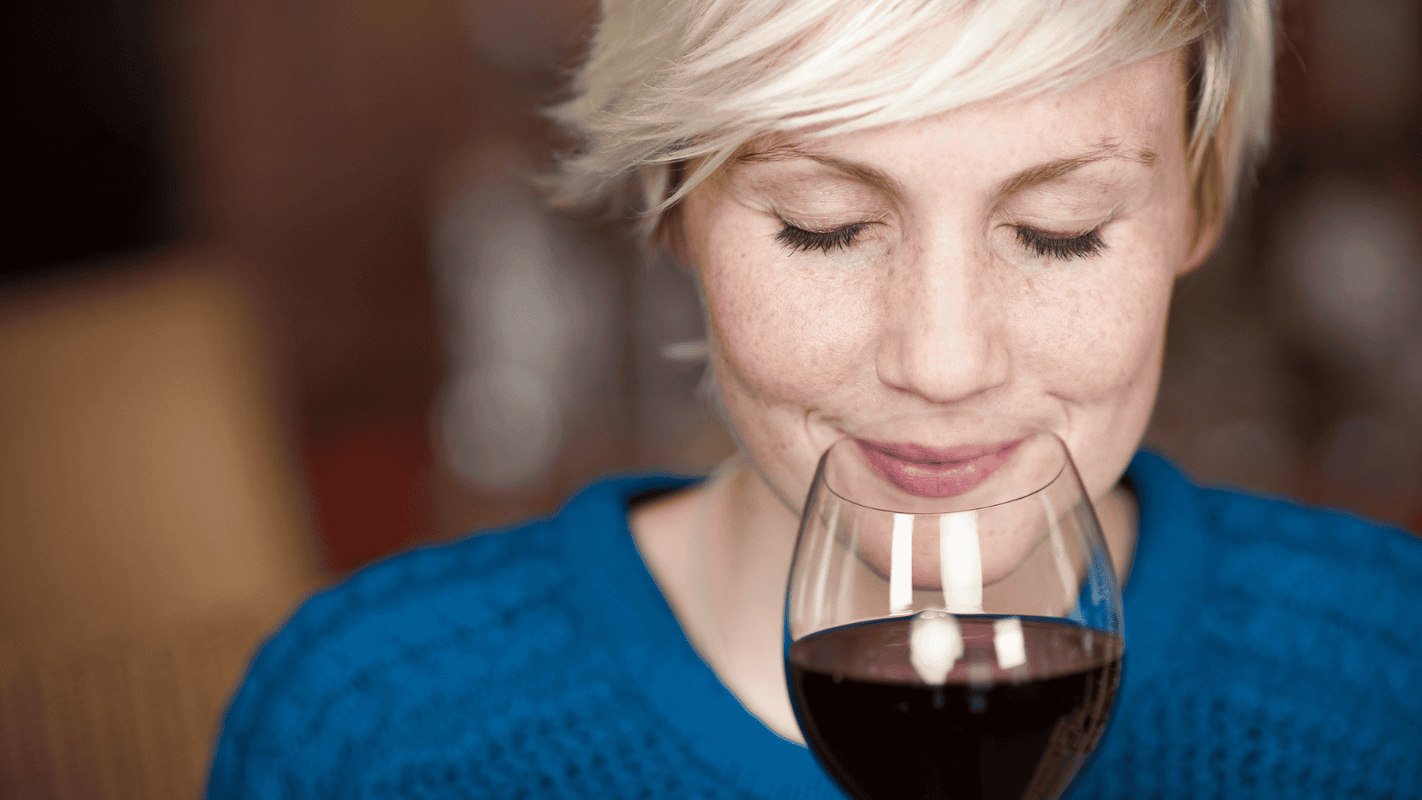 A person with short, blonde hair wearing a blue sweater gently holds a glass of red wine close to their nose with eyes closed, appearing to savor its aroma. The background is blurred.