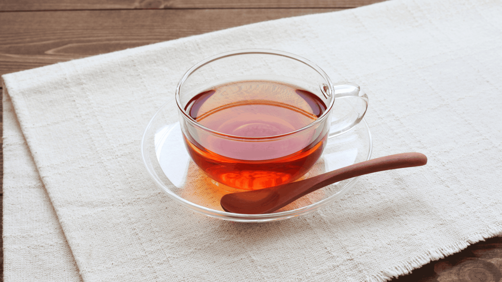 A glass cup filled with black tea is placed on a saucer atop a beige cloth. A wooden spoon rests on the saucer beside the cup. The setting is on a wooden surface.