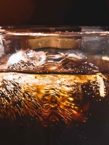 Close-up of a clear, fizzy soft drink in a glass with ice cubes, backlit by warm lighting, emphasizing the bubbles and texture of the drink.