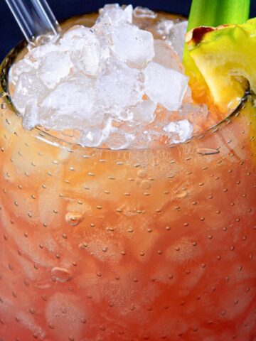 Close-up of a cold, dewy tropical drink in a textured glass, filled with crushed ice and garnished with a slice of pineapple and a green straw, set against a dark blue background.