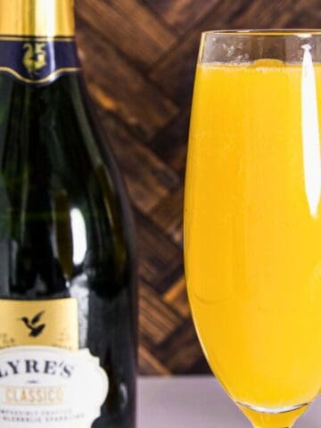 Two glasses filled with orange juice on a table next to a bottle labeled "lyre's classic," with a small potted plant and a patterned wooden backdrop.