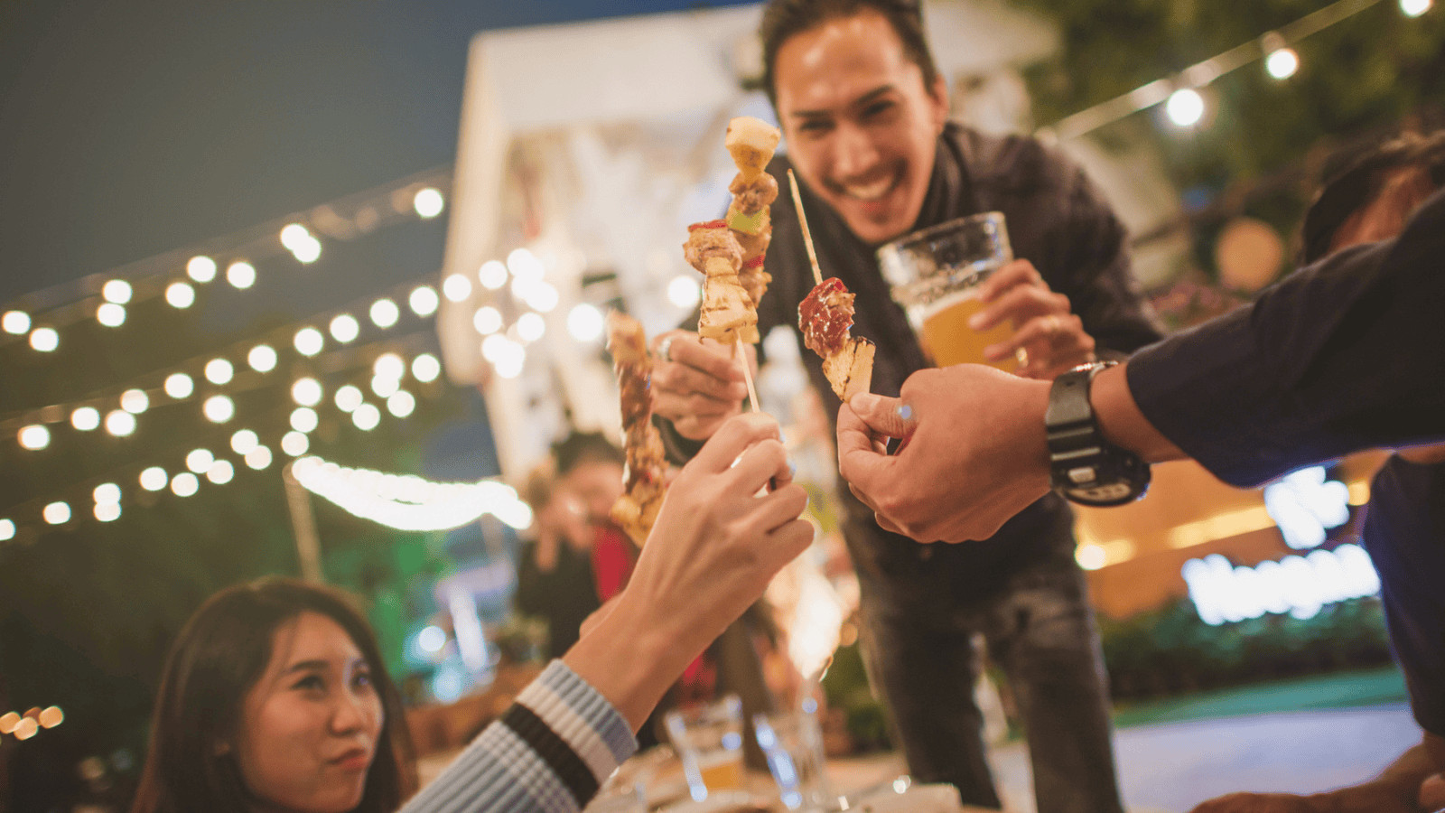 Group of people raising skewers in a toast while a man, holding a glass of beer, leans over in a festive outdoor setting decorated with string lights. One woman is visible at the left side of the table. The mood appears celebratory and cheerful.