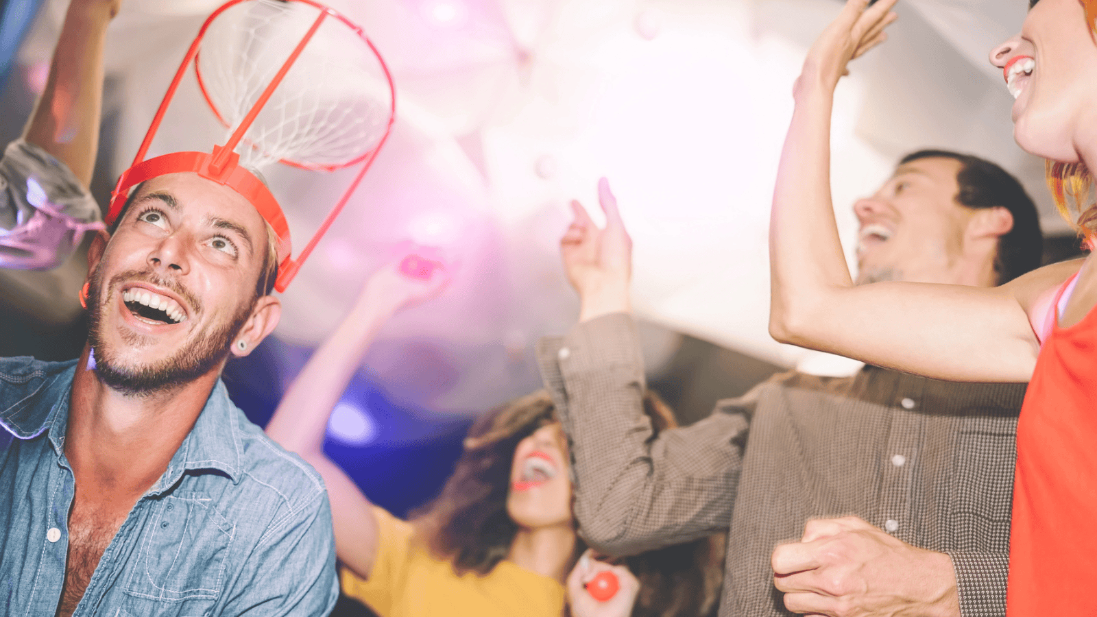 A group of people are dancing and having fun at a party. In the foreground, a man is wearing a plastic basketball hoop on his head and smiling. The background shows other people raising their arms and enjoying themselves in the brightly lit space.