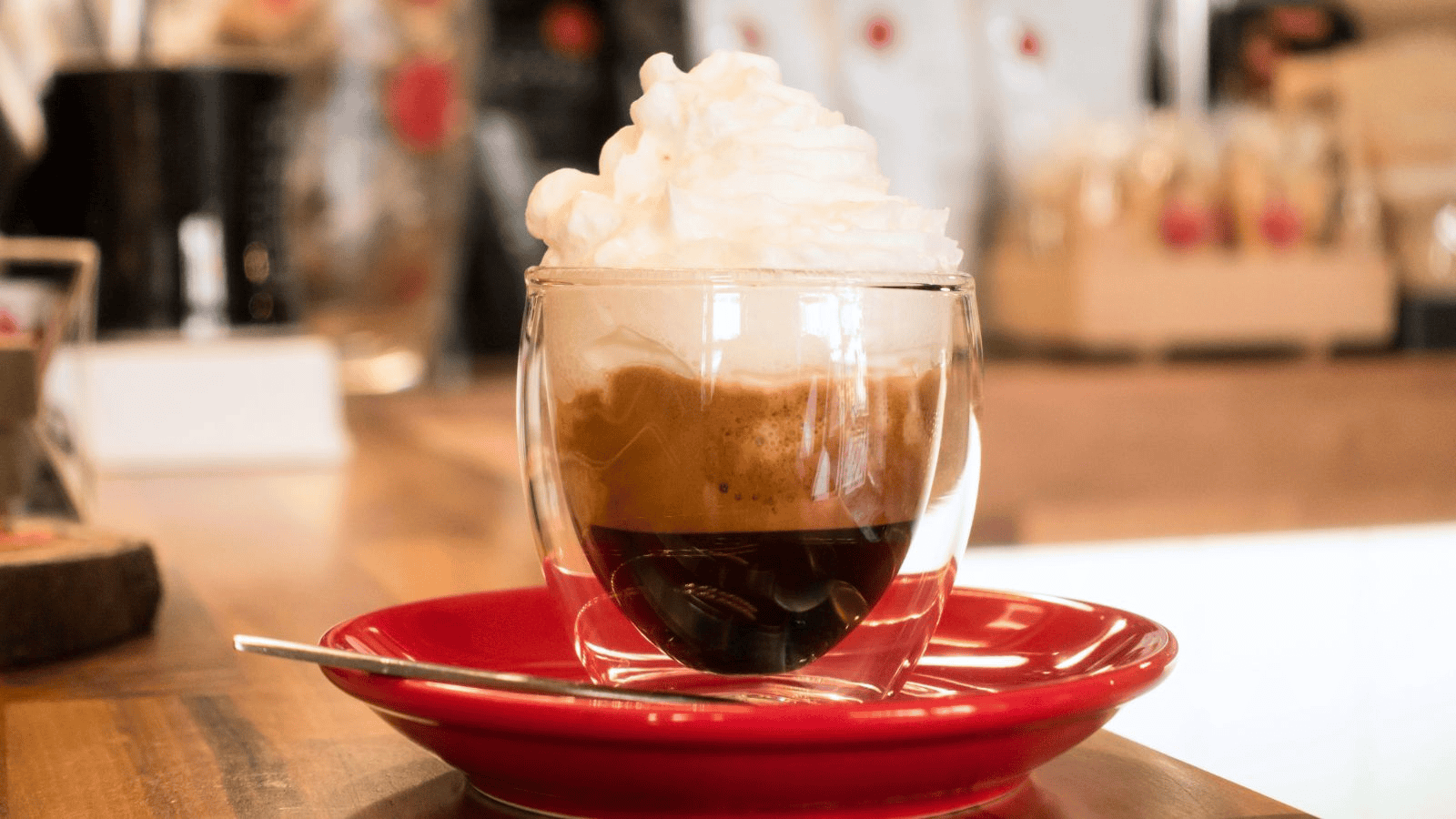 A small glass cup of coffee topped with a mound of whipped cream sits on a red saucer with a silver spoon. The background includes various blurry coffee shop elements.