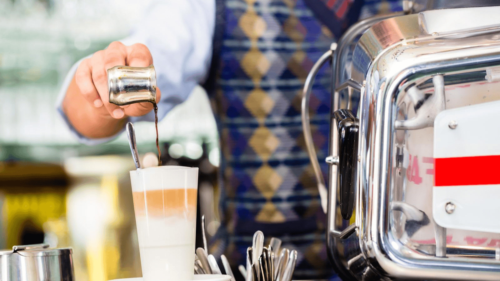 A person is pouring espresso from a small metal pitcher into a cup of layered coffee, positioned next to a coffee machine. The person is wearing a patterned vest and shirt sleeves are visible. Several spoons are placed in a holder nearby.