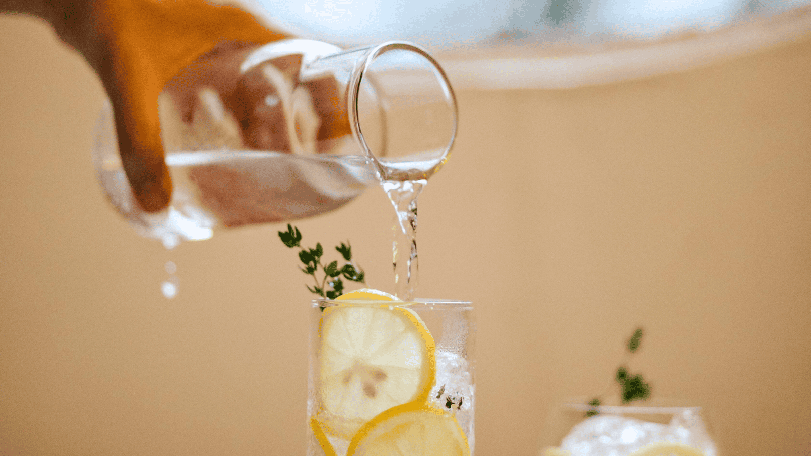 A hand pours lemon water from a glass carafe into a glass filled with ice cubes, lemon slices, and a sprig of thyme. The background is softly blurred, creating a warm, inviting atmosphere.