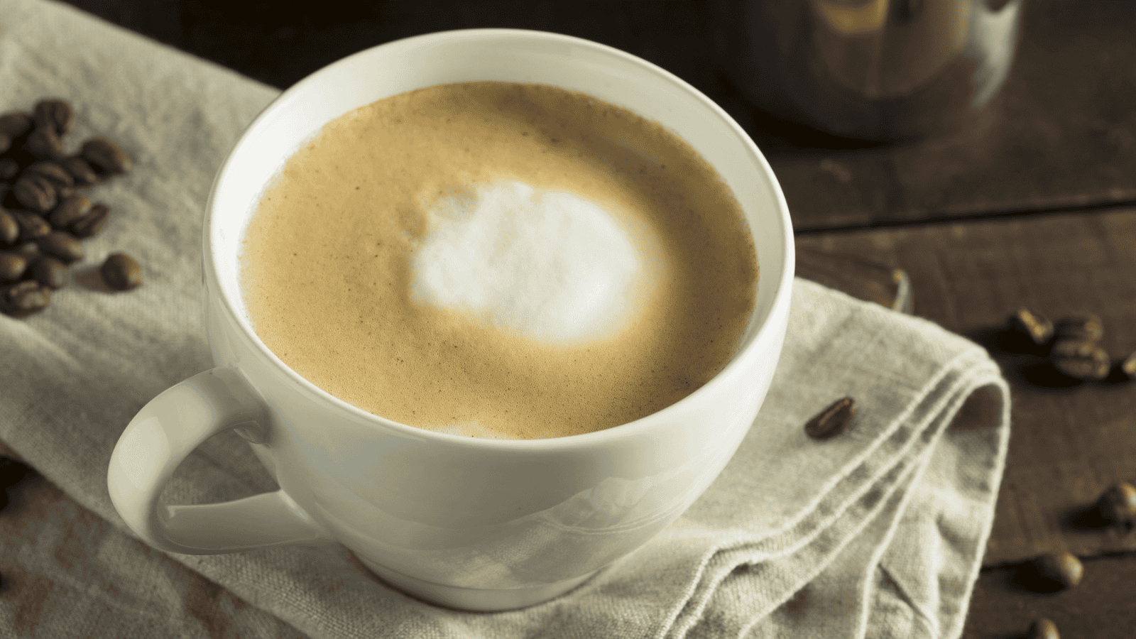 A white cup filled with a frothy cappuccino sits on a beige cloth napkin. Coffee beans are scattered around the napkin on a wooden surface. A metallic container is partially visible in the background.