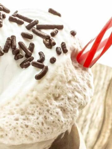 Close-up of a milkshake with whipped cream, chocolate sprinkles, and a cherry on top, served in a glass with a red and white striped straw.
