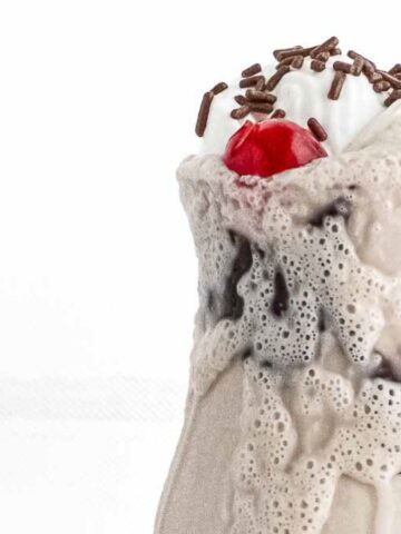 Milkshake overflowing its glass, topped with whipped cream, chocolate sprinkles, and a red cherry, served with a red and white striped straw, against a white background.