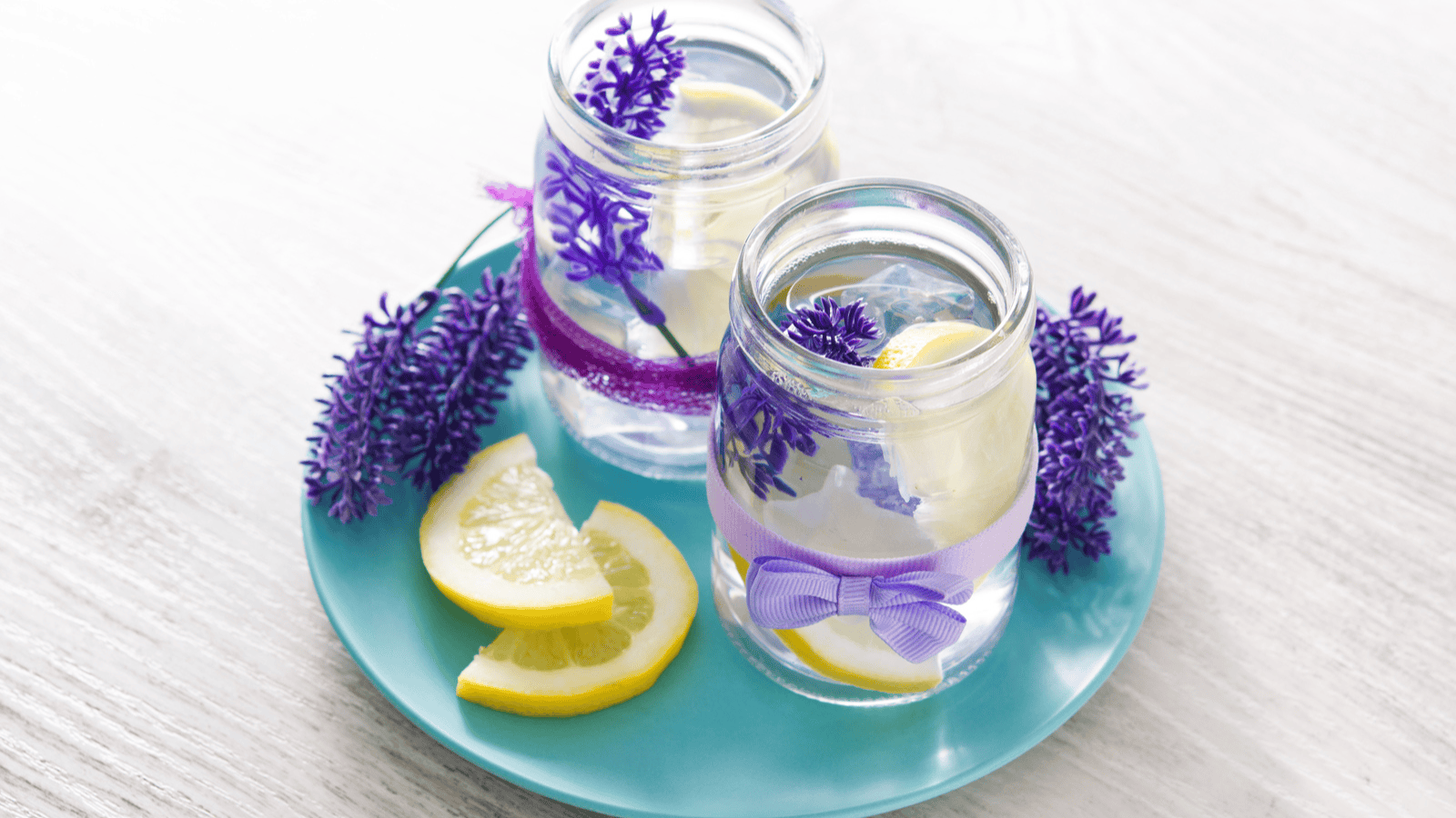 Two glass jars filled with clear liquid and lemon slices, decorated with purple flowers and tied with purple ribbons, are placed on a blue plate. There are also a few lemon slices and additional purple flowers on the plate.