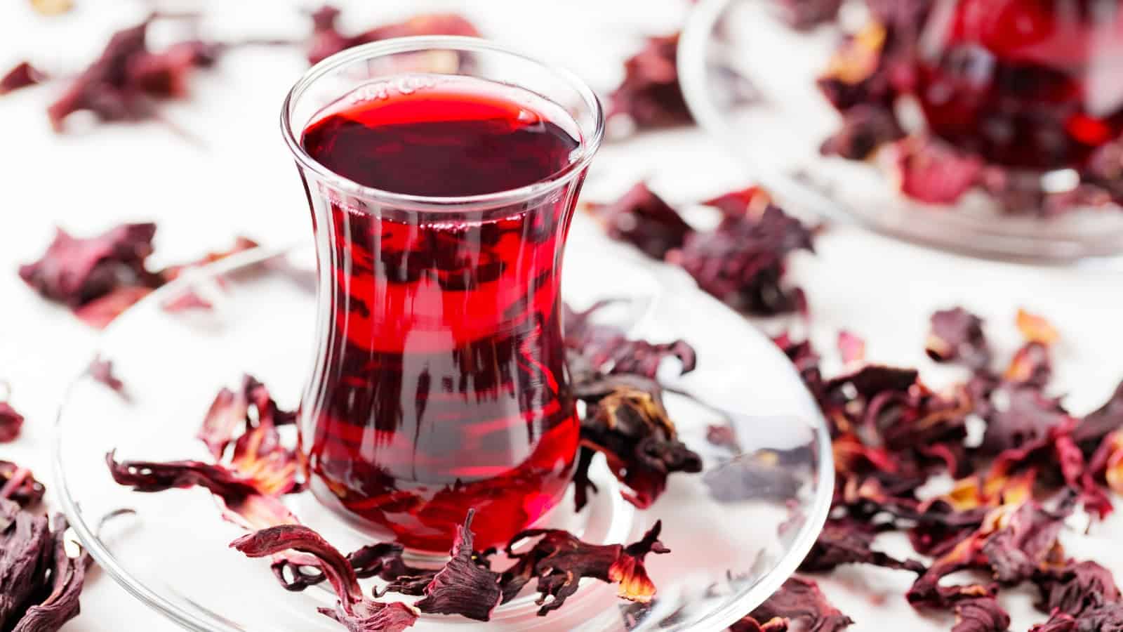 A clear glass filled with deep red hibiscus tea sits on a matching glass saucer. Dried hibiscus flowers are scattered around the glass and saucer on a white surface, adding an organic touch to the scene.