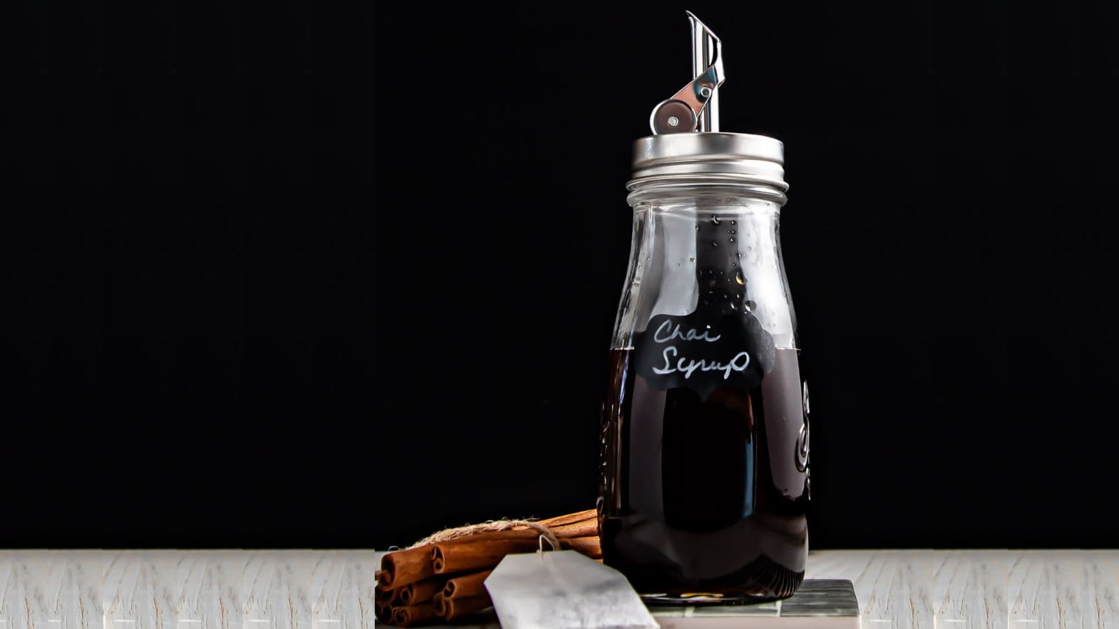 A glass jar labeled "chai syrup" filled with dark liquid, a pen nib as a decorative topper, next to cinnamon sticks and a white spoon, against a black background.