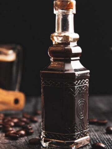 A darkly lit image featuring an ornate bottle of coffee liqueur with scattered coffee beans, a glass of liqueur, and a teapot in the background on a wooden surface.