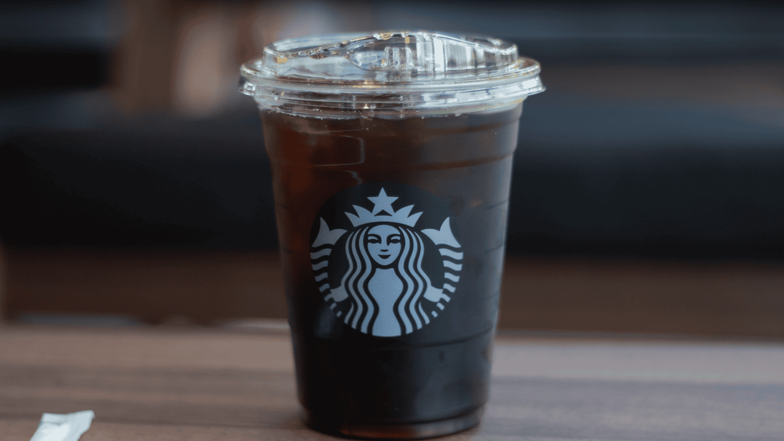 A clear plastic Starbucks cup filled with iced black coffee sits on a wooden table. The Starbucks logo is prominently displayed on the front of the cup.