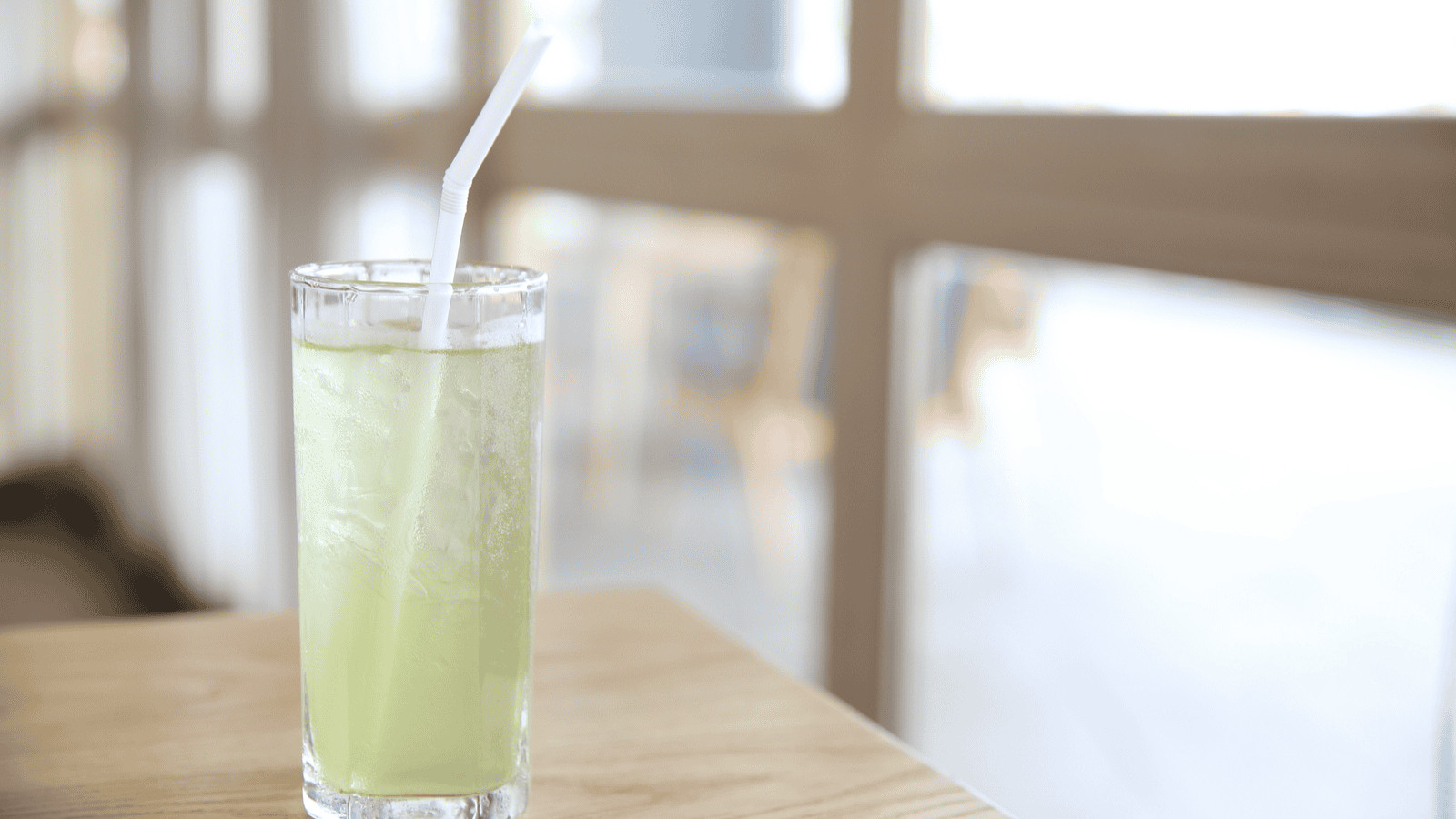 A clear glass filled with a light green beverage, topped with ice and a white straw, is placed on a wooden table. The background shows a blurred indoor setting with windows and chairs.