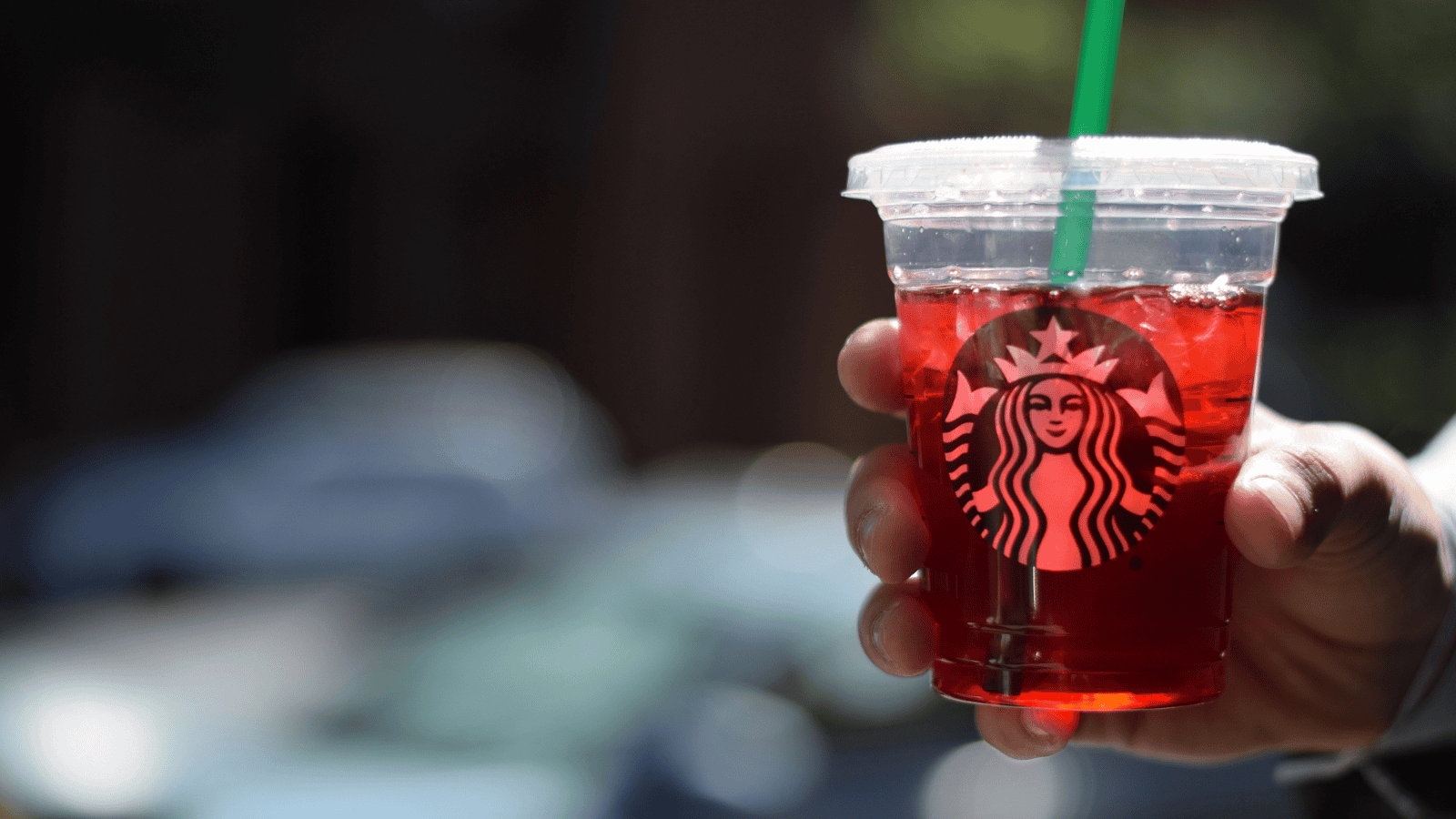 A hand holds a clear plastic cup with a green straw, filled with a red iced beverage—a sugar-free Starbucks drink—featuring the iconic logo. The background is blurred, hinting at an outdoor setting with dim lighting.