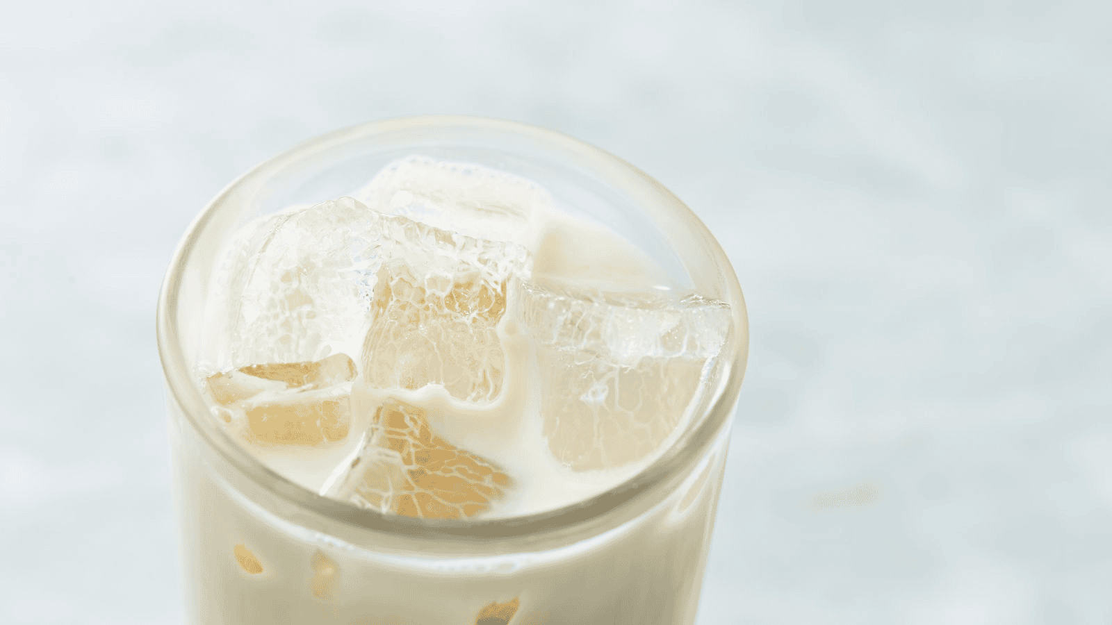A close-up of a glass filled with a creamy beverage, likely iced milk tea or a similar drink, showing large ice cubes floating in the liquid. The background is softly blurred, with a light, neutral color.