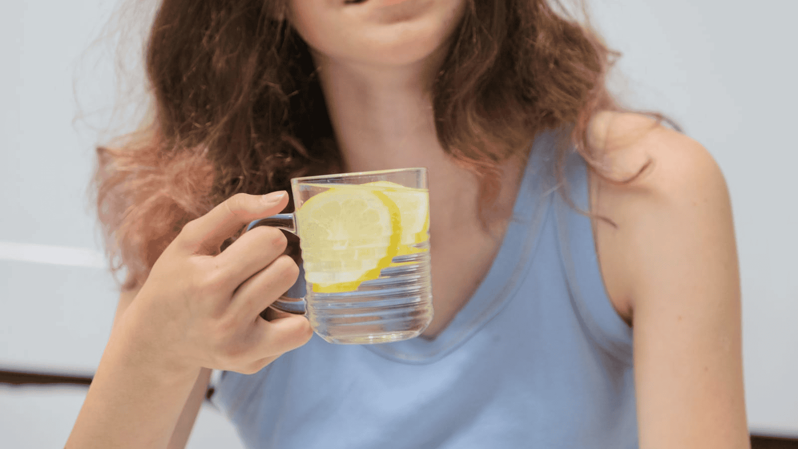 A person with long, wavy hair and wearing a light blue tank top holds a glass of water with lemon slices. The setting appears indoor with a light-colored background. The person's face is partially visible.
