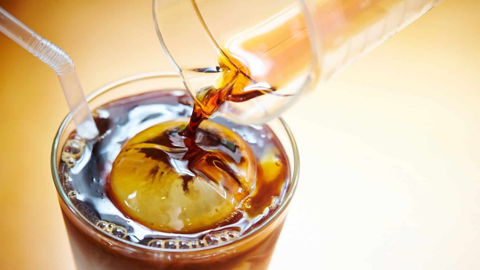 A close-up image of iced coffee being poured into a glass with ice cubes and a straw. The liquid is captured mid-pour, creating a dynamic motion as it circulates around the ice. The background is blurred and warm in tone.