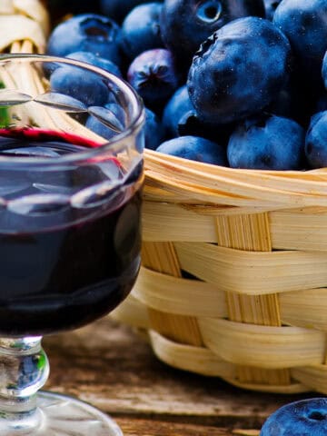 A glass of blueberry juice is placed on a wooden surface next to a wicker basket filled with fresh blueberries. Some green leaves are scattered around the basket.